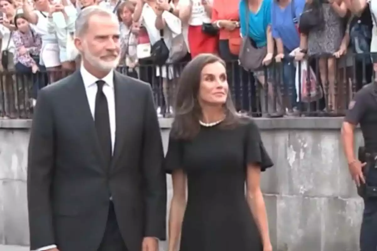 Una pareja elegantemente vestida de negro se encuentra de pie frente a una multitud que los observa y toma fotos.