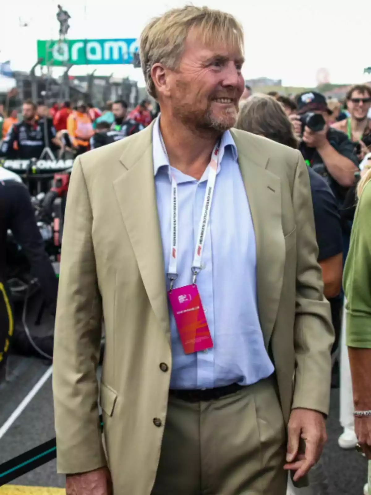 El rey Guillermo de Holanda con traje beige y camisa azul claro lleva una acreditación colgada al cuello mientras sonríe en un evento al aire libre rodeado de personas y fotógrafos. El rey Guillermo de Holanda con traje beige y camisa azul claro lleva una acreditación colgada al cuello mientras sonríe en un evento al aire libre rodeado de personas y fotógrafos.