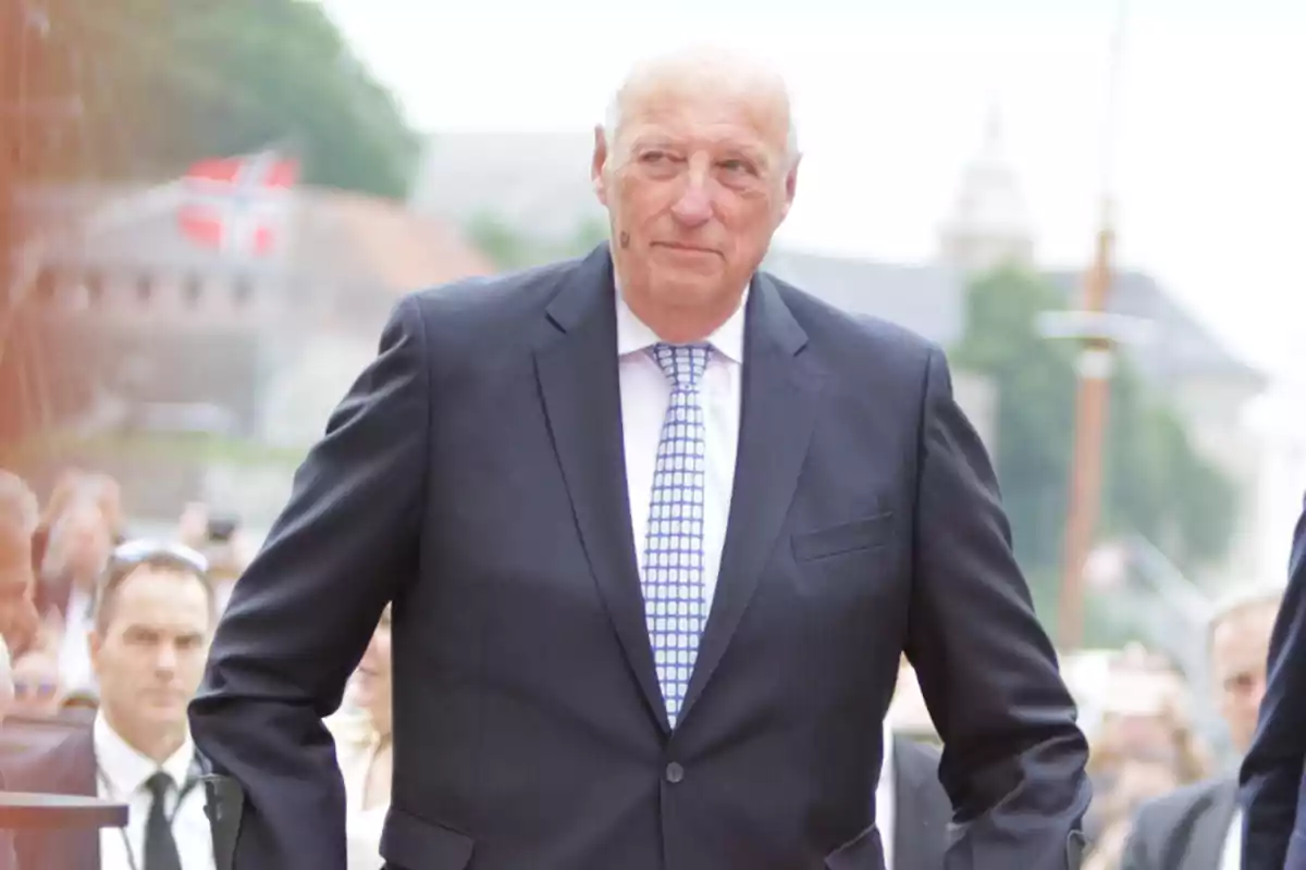 El rey Harald de Noruega con traje oscuro y corbata de cuadros, de pie al aire libre con una bandera noruega de fondo. El rey Harald de Noruega con traje oscuro y corbata de cuadros, de pie al aire libre con una bandera noruega de fondo.