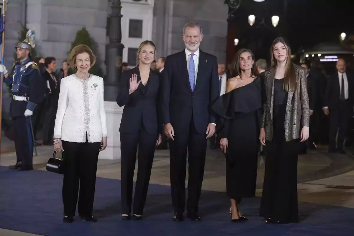 Un grupo de personas elegantemente vestidas posando en un evento nocturno con un fondo de guardias y luces. Un grupo de personas elegantemente vestidas posando en un evento nocturno con un fondo de guardias y luces.
