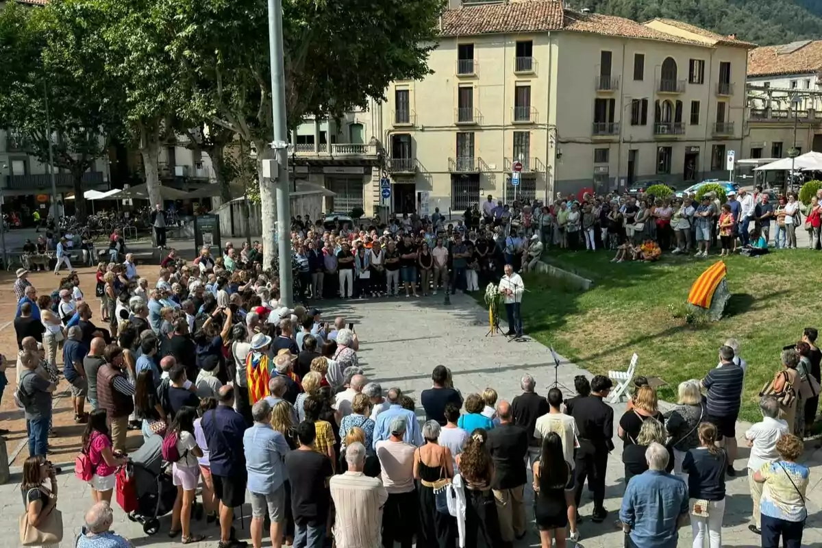 Una multitud de persones s'han reunit en una plaça a l'aire lliure per escoltar un orador que està dret davant d'un micròfon i un ram de flors mentre un edifici de diversos pisos i arbres grans es troben al fons.