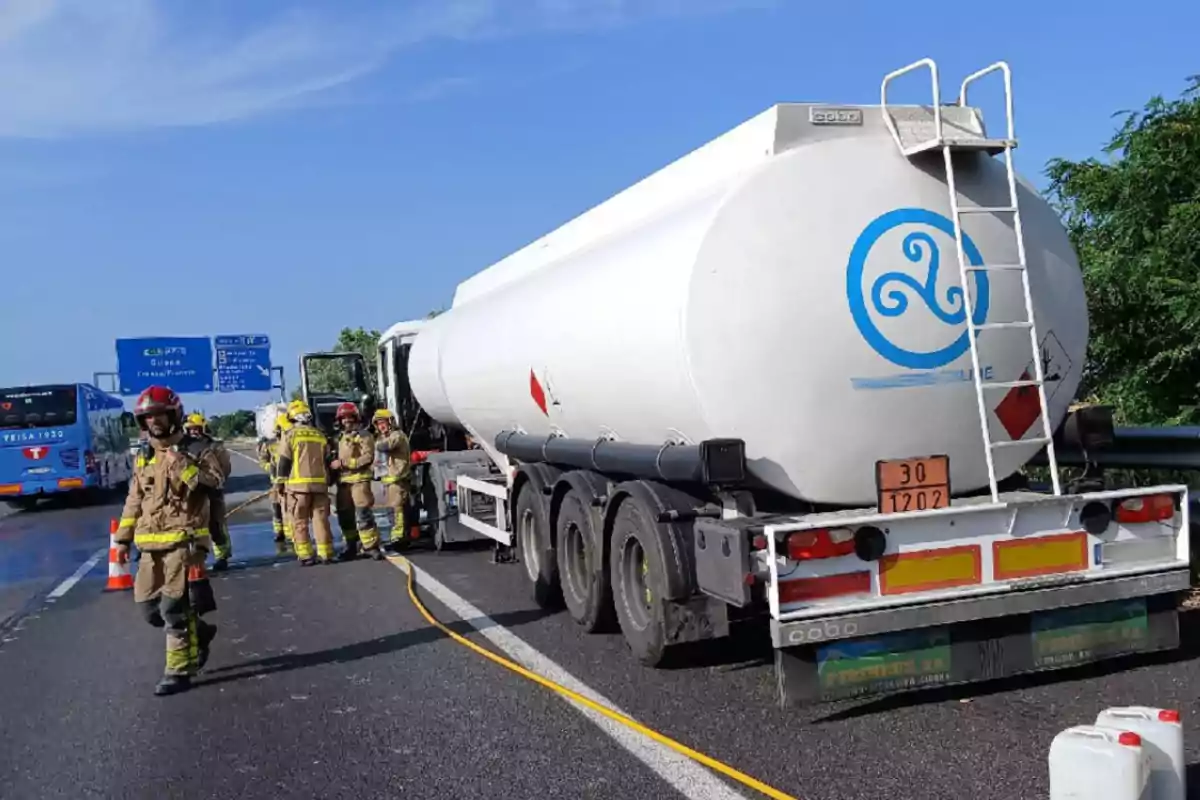 Un camió cisterna aturat en una carretera amb diversos bombers treballant al seu voltant i un autobús blau al fons