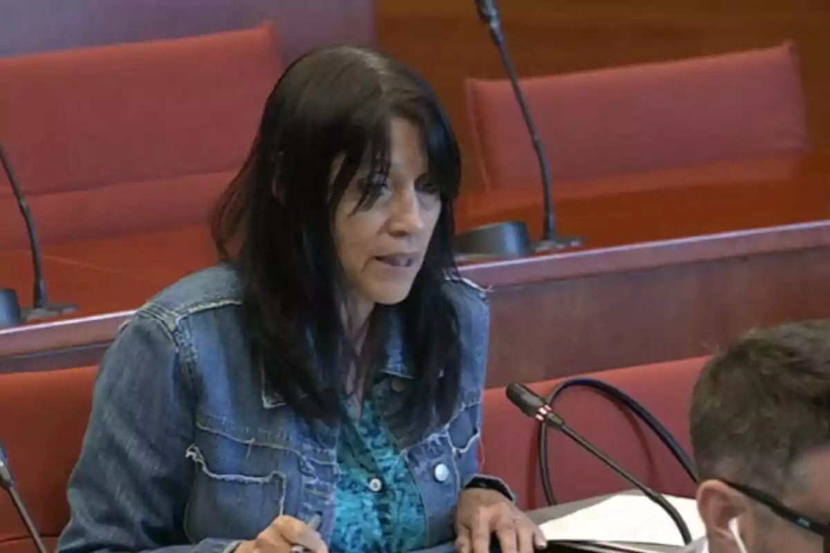 Dona de cabell fosc parlant davant d’un micròfon en una sala de conferències amb seients vermells