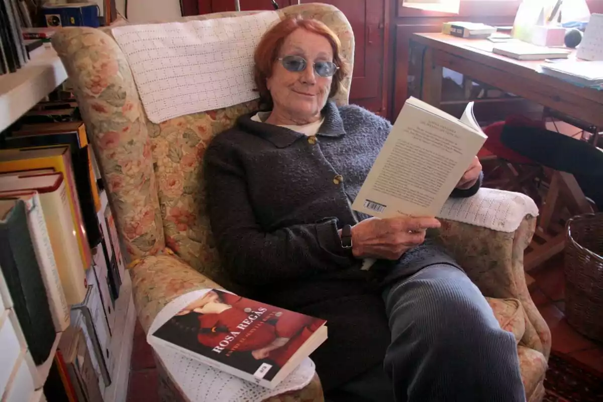 Una dona gran amb ulleres blaves està asseguda en una butaca floral llegint un llibre, amb un altre llibre titulat 