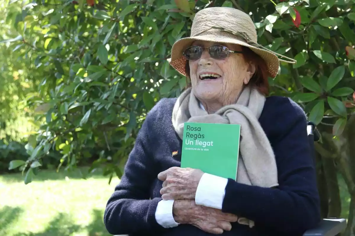 Una dona gran somriu mentre sosté un llibre verd titulat "Rosa Regàs Un arribat" en un jardí amb vegetació abundant.