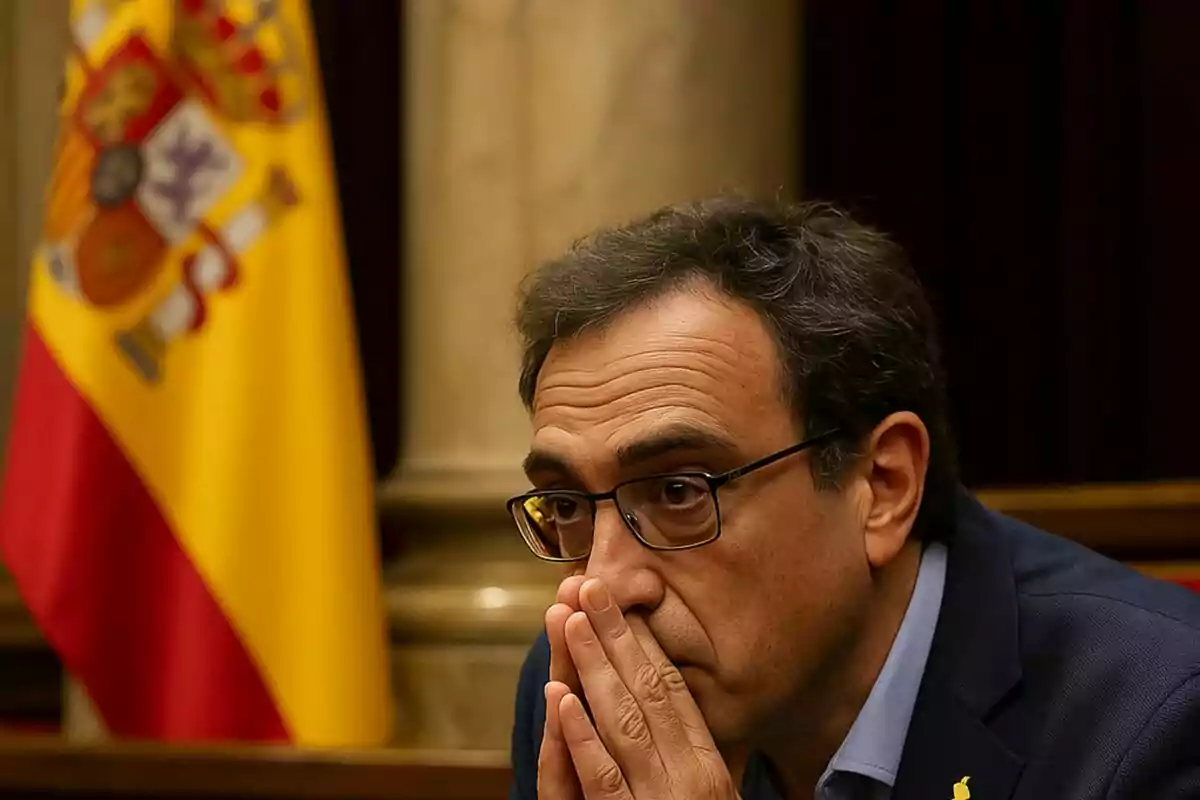 Un home amb ulleres i expressió pensativa es troba en un entorn formal amb una bandera d'Espanya al fons.