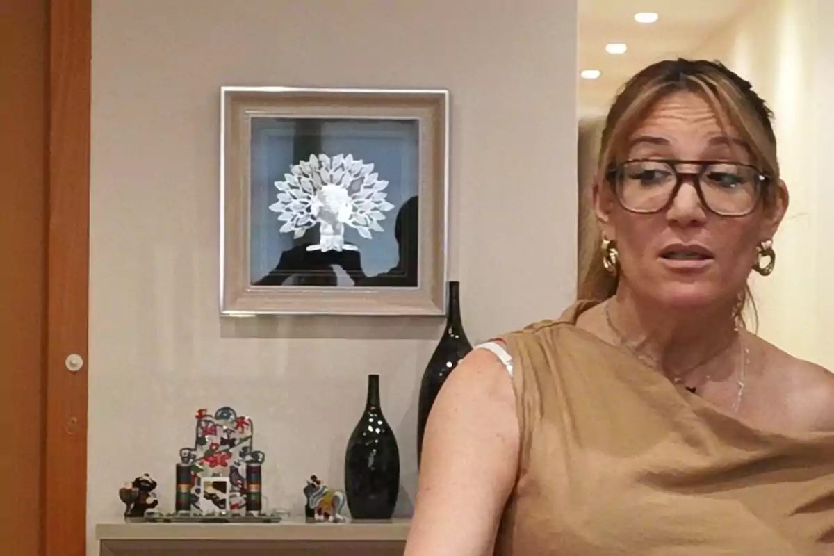 Una dona amb ulleres i arracades daurades està dreta davant d'una paret decorada amb un quadre d'art i diverses figures. Una dona amb ulleres i arracades daurades està dreta davant d'una paret decorada amb un quadre d'art i diverses figures.