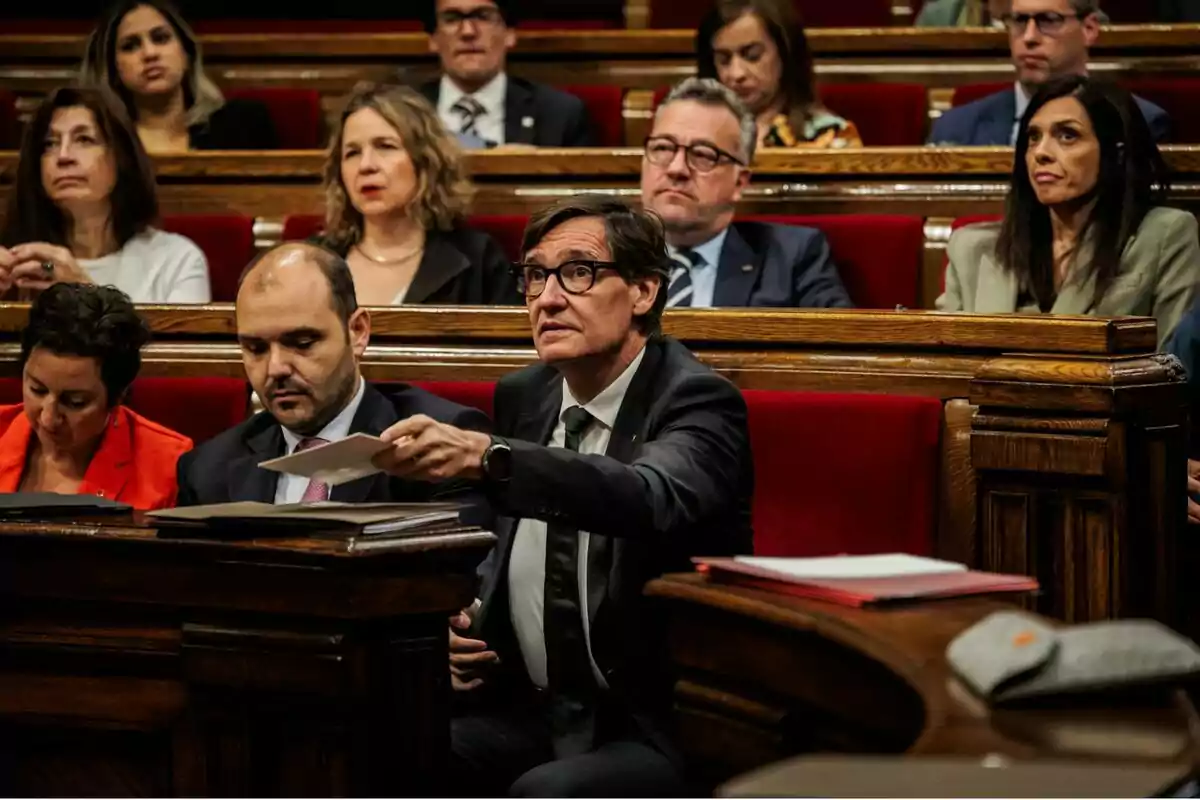 Un grup de persones assegudes en un parlament, algunes d'elles prestant atenció mentre un home al centre sosté un paper.