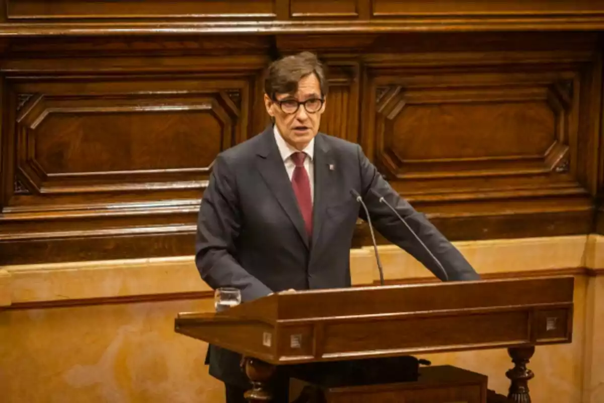 Hombre con gafas y traje oscuro hablando en un podio de madera en un entorno formal.