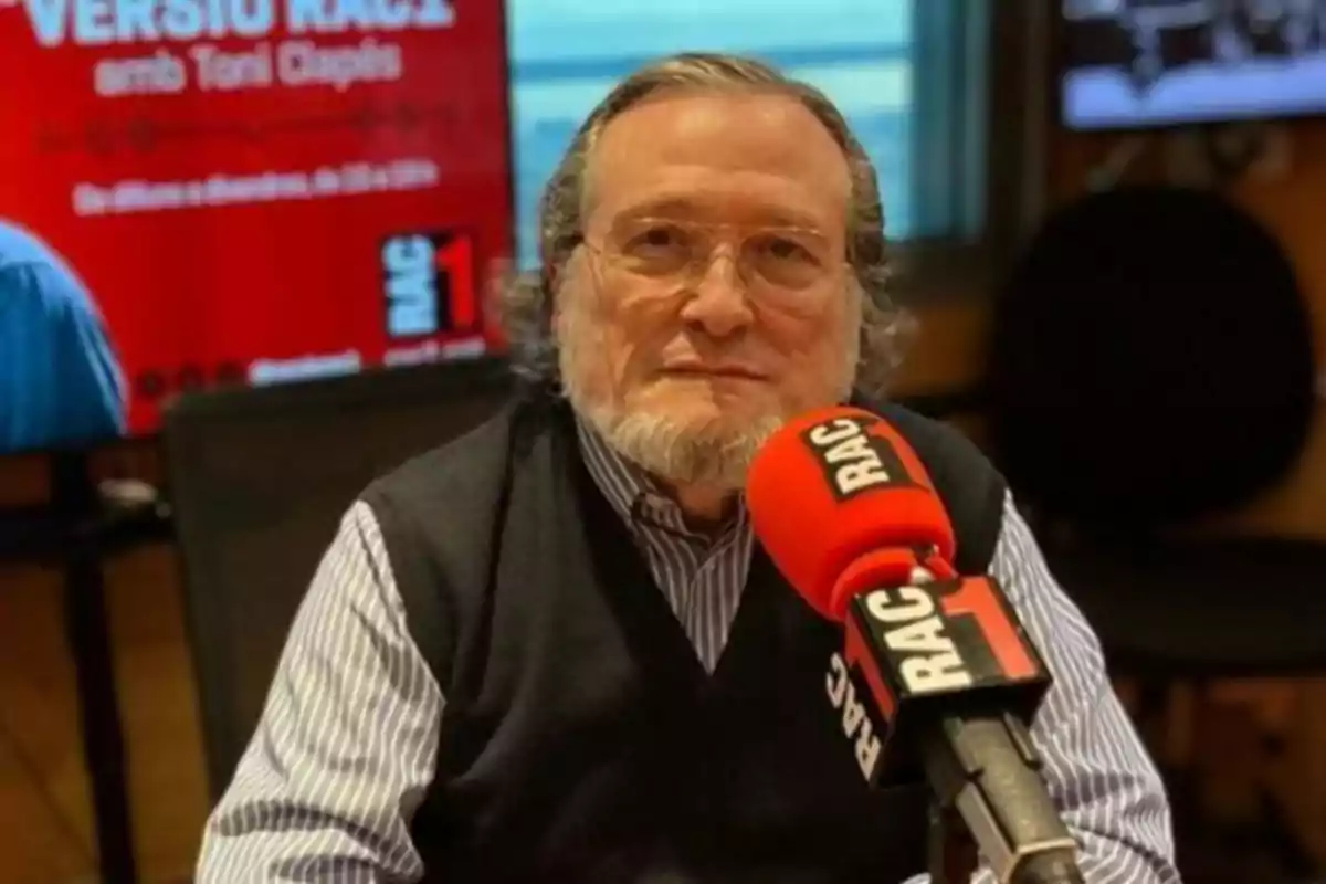 Un home gran amb ulleres i barba està assegut davant d'un micròfon vermell de RAC1 en un estudi de ràdio, amb un cartell vermell de fons que diu “VERSIÓ RAC1 amb Toni Clapés”.