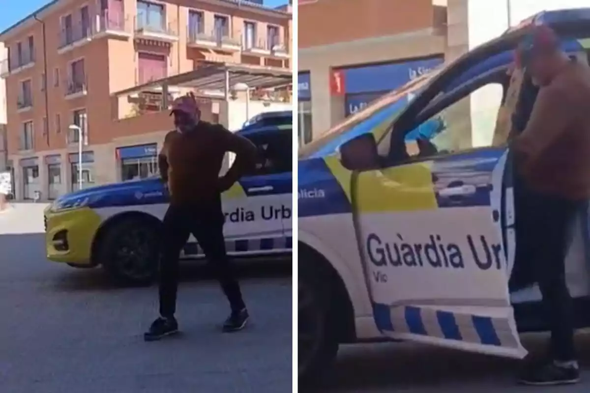 Un hombre con gorra y mascarilla está de pie junto a un coche de la Guardia Urbana en una calle con edificios de fondo.