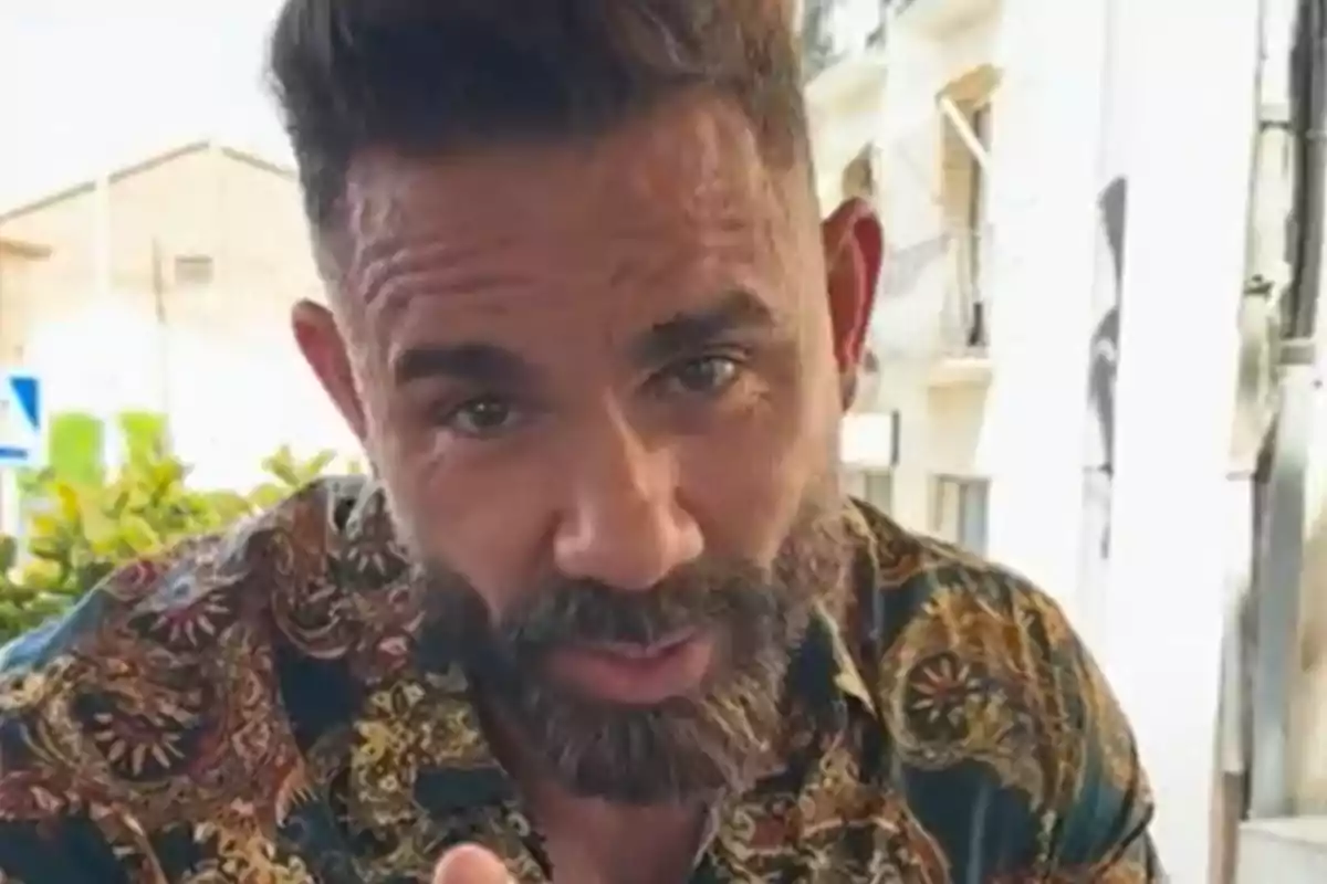 Un home amb barba i cabell curt, vestint una camisa de color estampat, mira directament a la càmera amb una expressió seriosa.