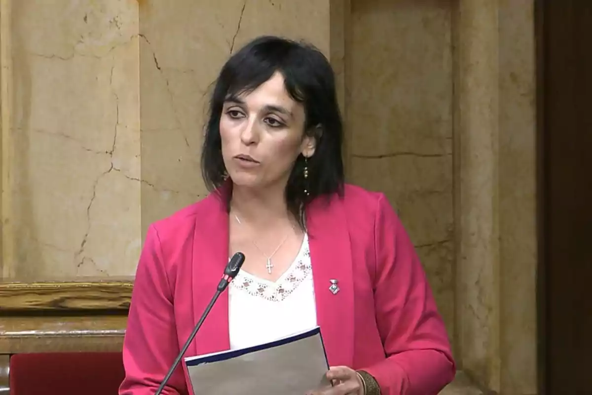 Dona de cabell fosc parlant en un podi amb un micròfon i sostenint papers, vestida amb jaqueta rosa i brusa blanca.