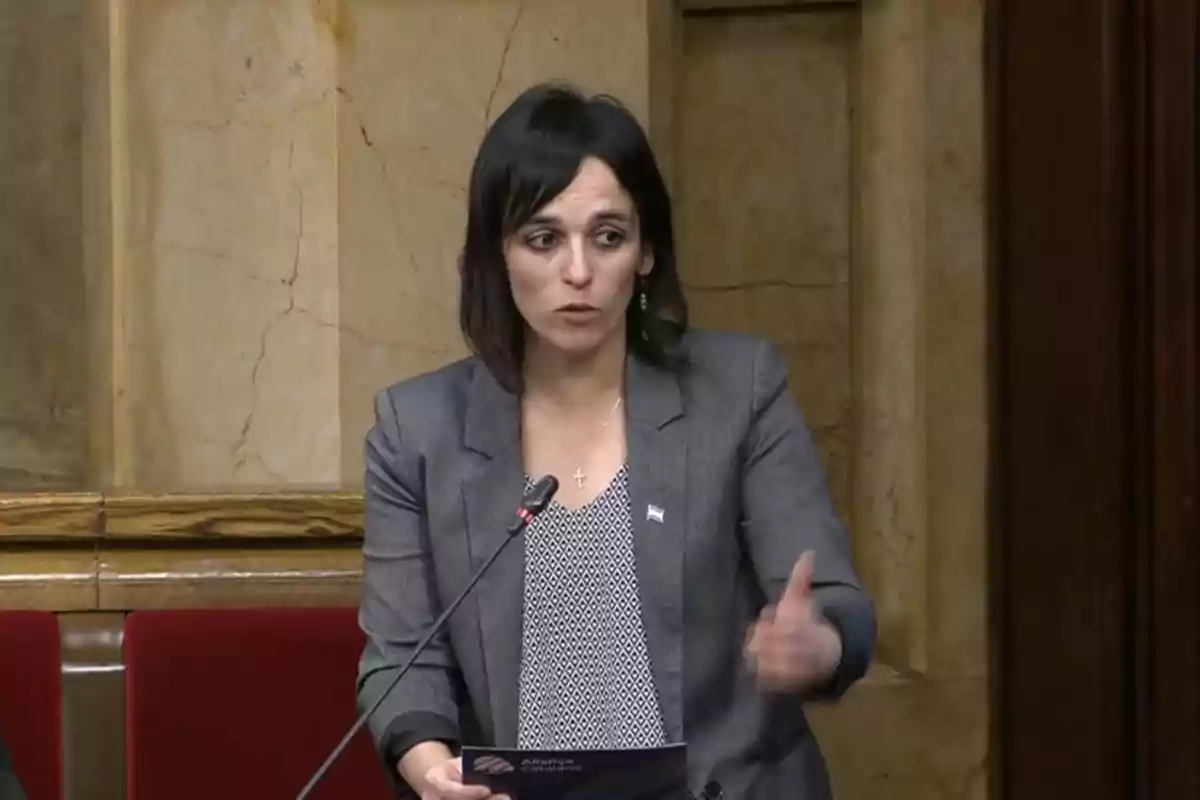 Una dona de cabell fosc i vestimenta formal parla en un podi en un entorn interior amb parets de marbre.