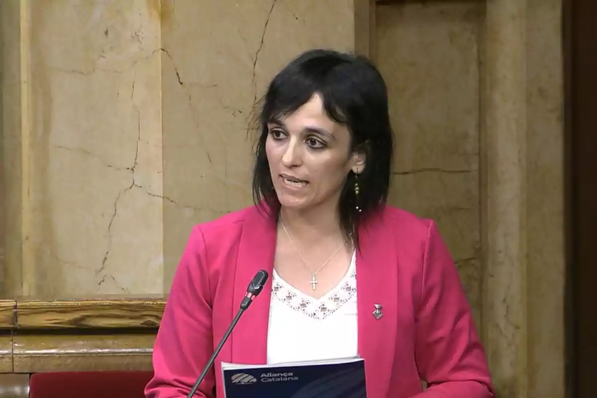 Una persona hablando en un podio con un fondo de mármol, vistiendo un blazer rosa y sosteniendo un documento.