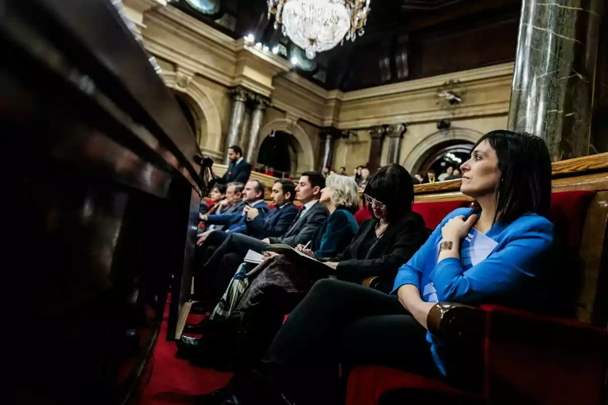 Persones assegudes en un auditori elegant amb columnes i un gran canelobre, algunes prenent notes i altres observant atentament.