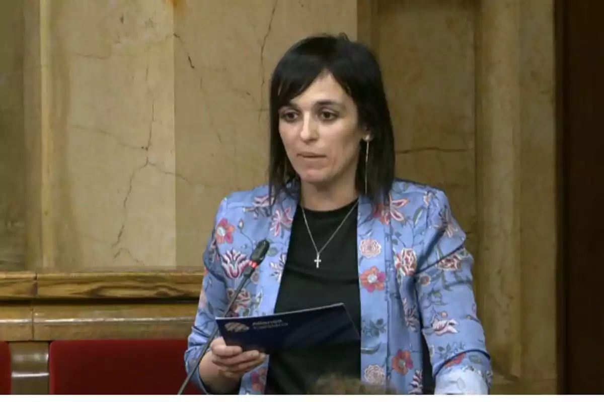 Una dona amb jaqueta blava de flors parla en un entorn formal mentre sosté un document.