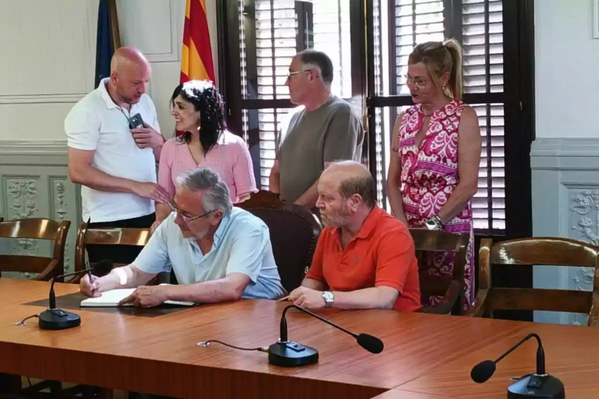Sis persones en una sala de reunions, dues d'elles assegudes a una taula de fusta amb micròfons, mentre una signa un document i les altres observen dretes al fons.