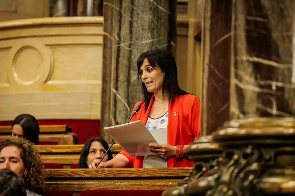 Dona de cabells foscos i jaqueta vermella parlant en un parlament mentre sosté uns papers