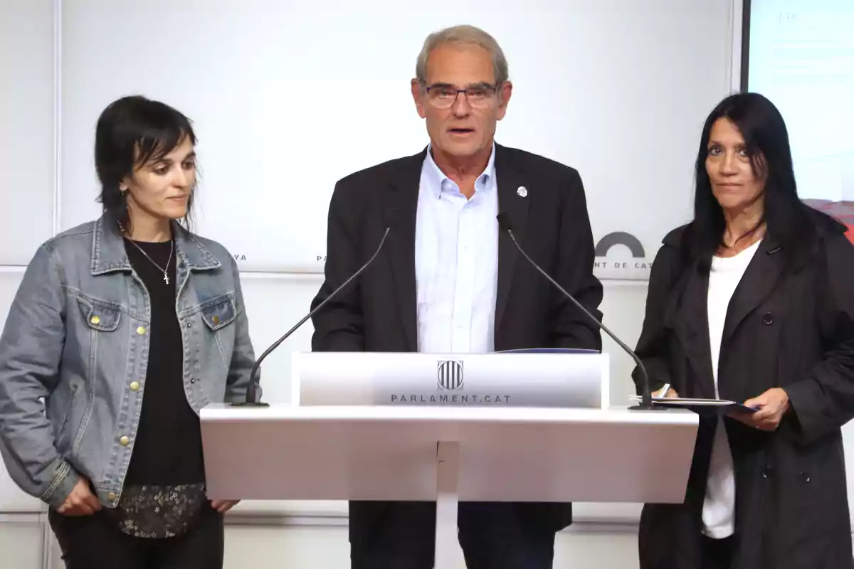 Tres persones dempeus en un podi amb el logotip del Parlament de Catalunya, una dona amb jaqueta de mezclilla a l'esquerra, un home amb ulleres i vestit al centre, i una dona amb jaqueta negra a la dreta.