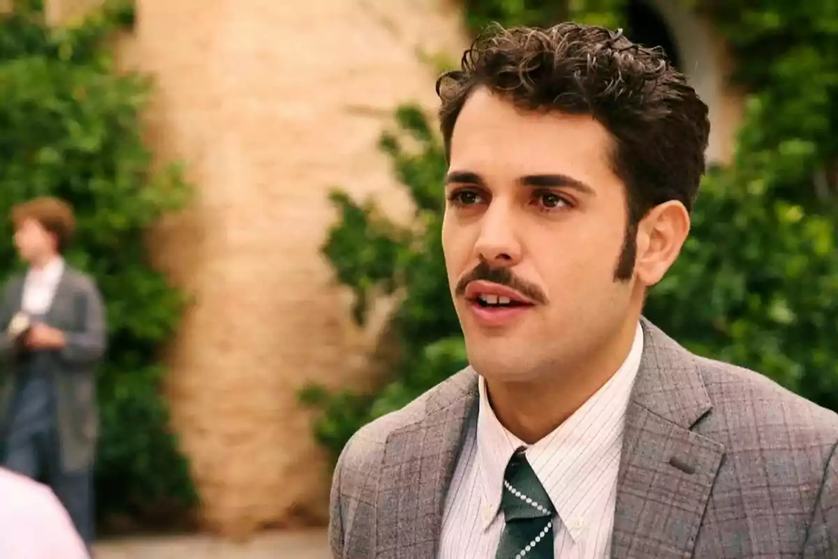 Santiago con bigote y traje a cuadros está al aire libre con un fondo de vegetación y una pared de ladrillo. Captura de 'Sueños de libertad' Santiago con bigote y traje a cuadros está al aire libre con un fondo de vegetación y una pared de ladrillo. Captura de 'Sueños de libertad'