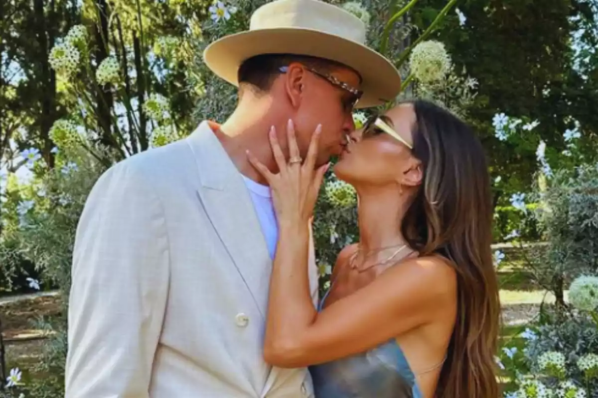 Una pareja besándose en un jardín con flores y árboles al fondo.