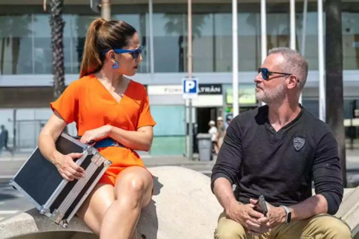 Una dona amb vestit taronja i ulleres de sol blaves sosté una capsa mentre conversa amb un home de barba canosa i ulleres de sol, tots dos asseguts a l'aire lliure davant d'un edifici modern.