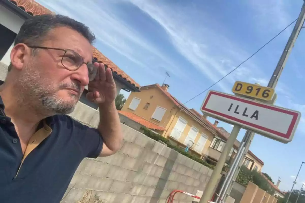 Un home amb ulleres i barba saluda militarment al costat d'un cartell de carretera que diu “ILLA” en una zona residencial amb cases de sostre vermell i cel clar.