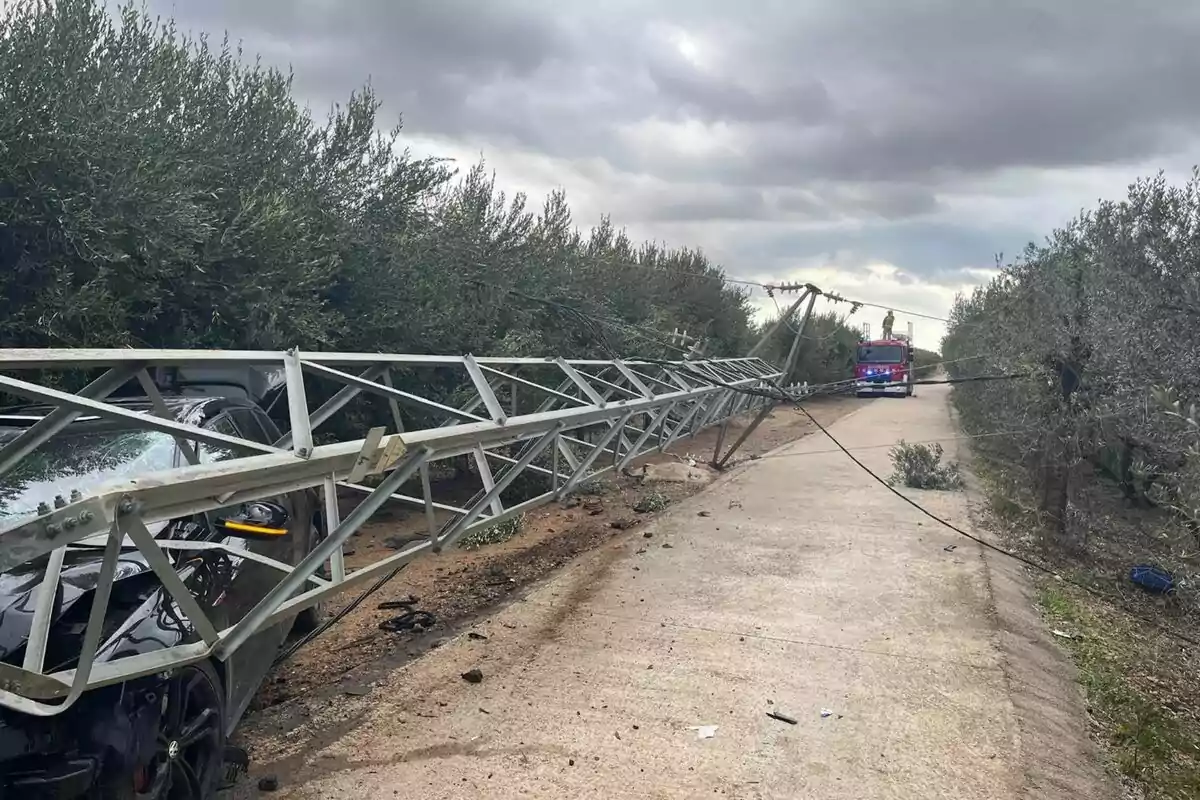 Un cotxe xoca contra una torre elèctrica que ha caigut sobre un camí envoltat d'arbres mentre un camió de bombers s'aproxima al lloc sota un cel ennuvolat.