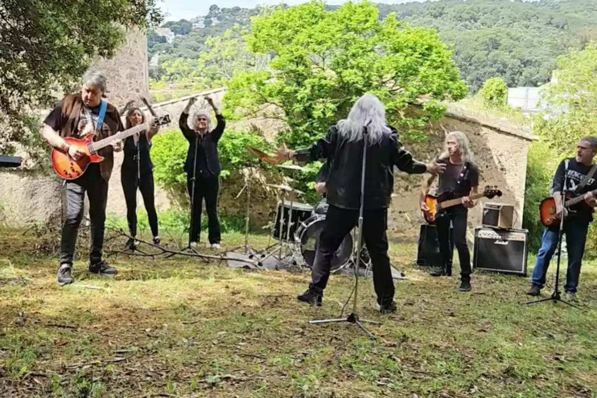 Un grup de músics estan tocant a l'aire lliure en un entorn natural, amb arbres i turons al fons, mentre algunes persones aplaudeixen i gaudeixen de la música.