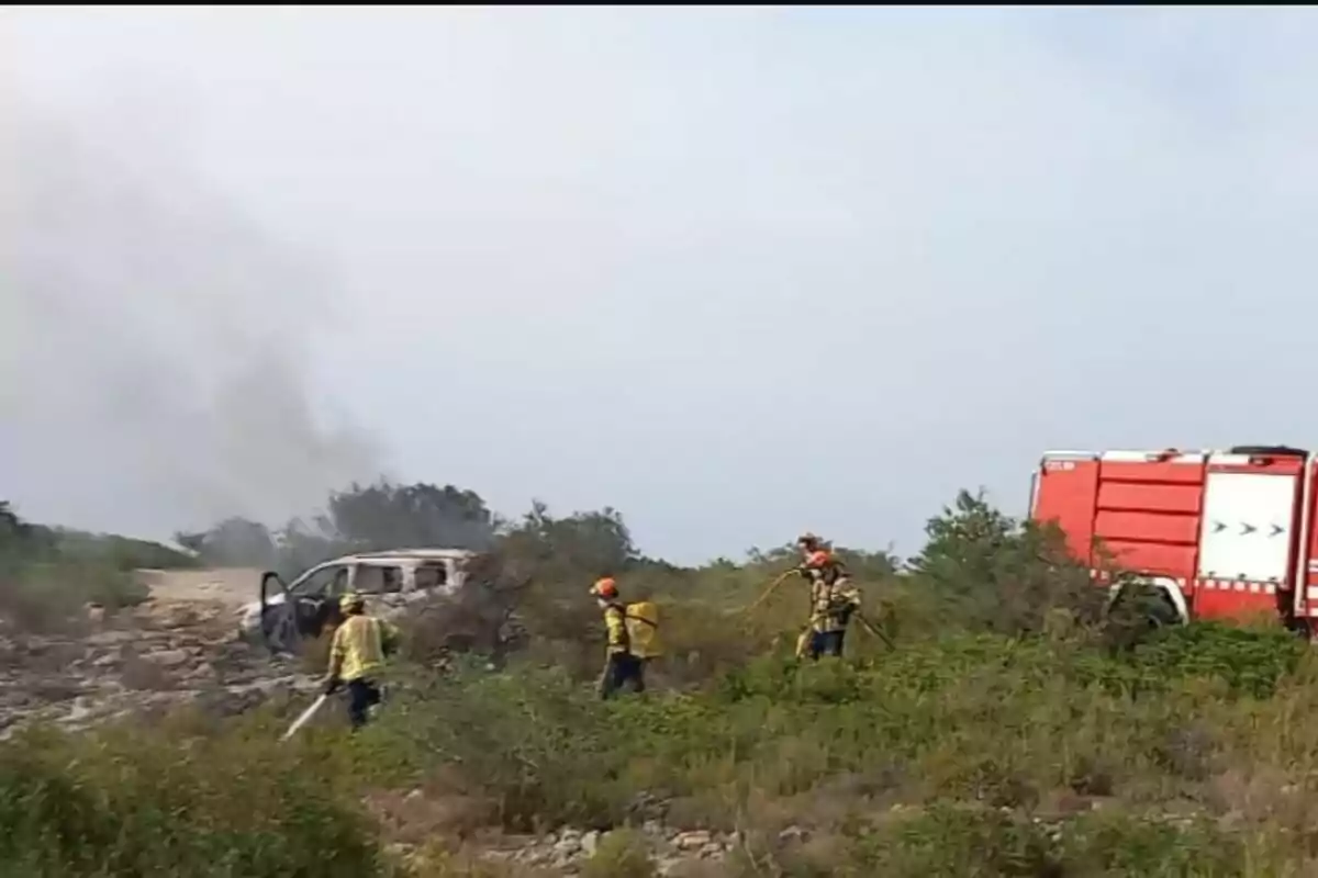 Bombers amb equip de protecció treballen en una zona de pastures on s’observa un vehicle cremat i un camió de bombers vermell.