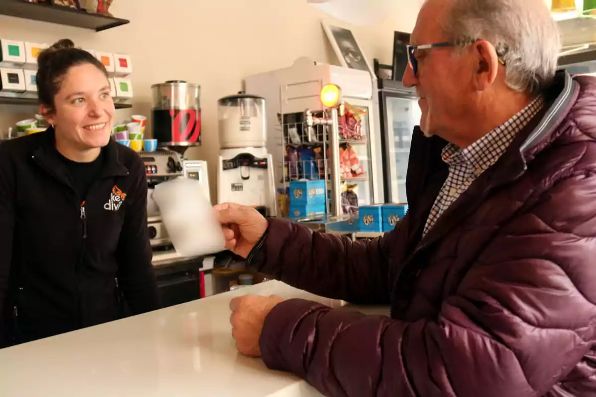 Un home gran amb ulleres i jaqueta morada sosté una tassa mentre conversa amb una dona darrere del taulell en una cafeteria.