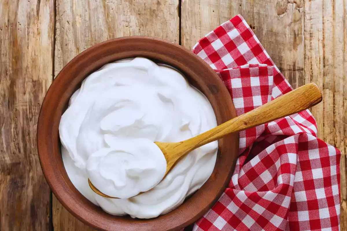 Un bol de ceràmica amb crema blanca i una cullera de fusta sobre una taula de fusta amb unes estovalles a quadres vermells i blancs.