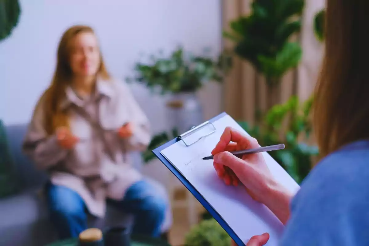 Una persona pren notes en un porta-retalls mentre una altra persona està asseguda al fons en un entorn amb plantes.