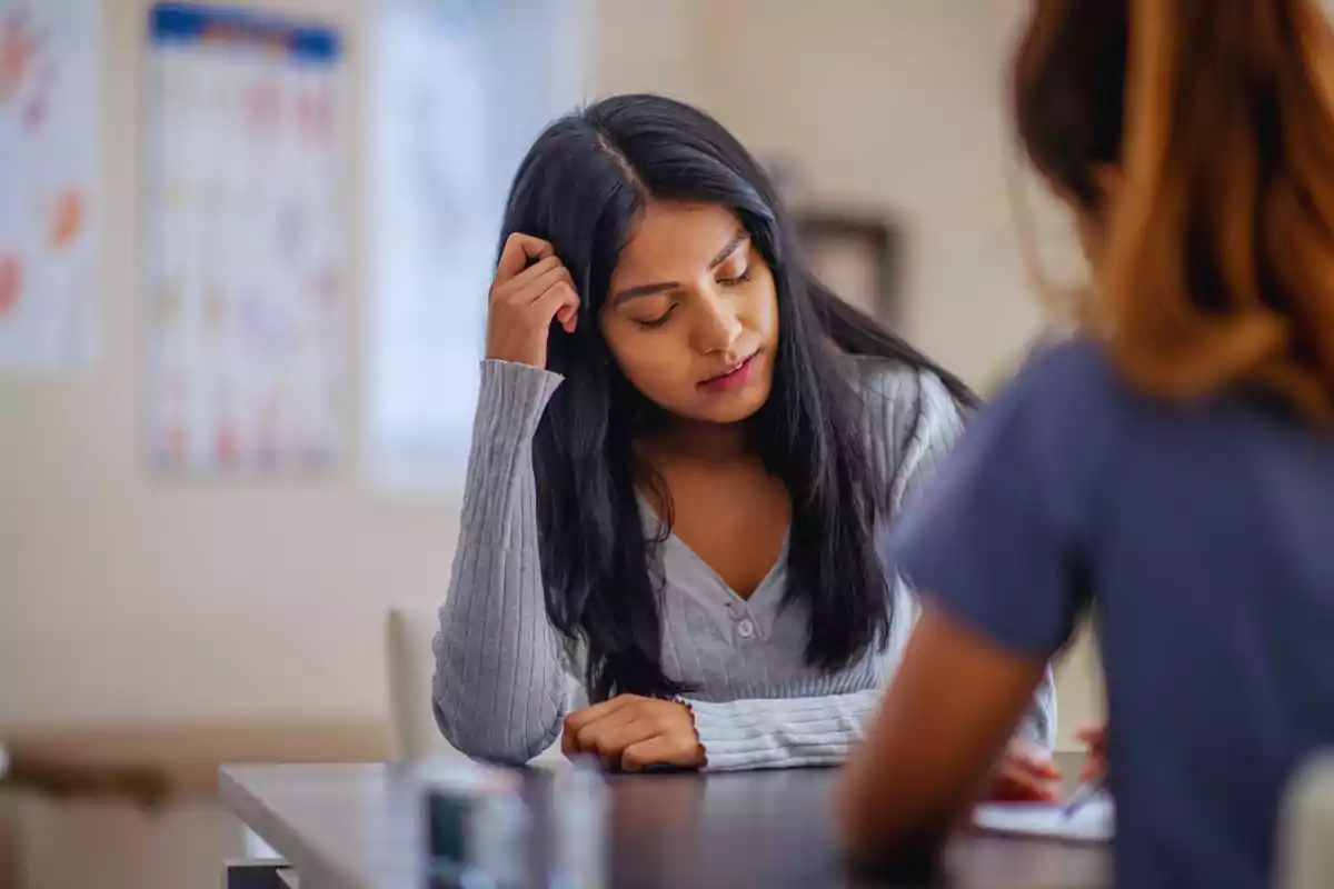 Una mujer joven con cabello largo y oscuro está sentada en una mesa, mirando hacia abajo con expresión pensativa, mientras otra persona está frente a ella.