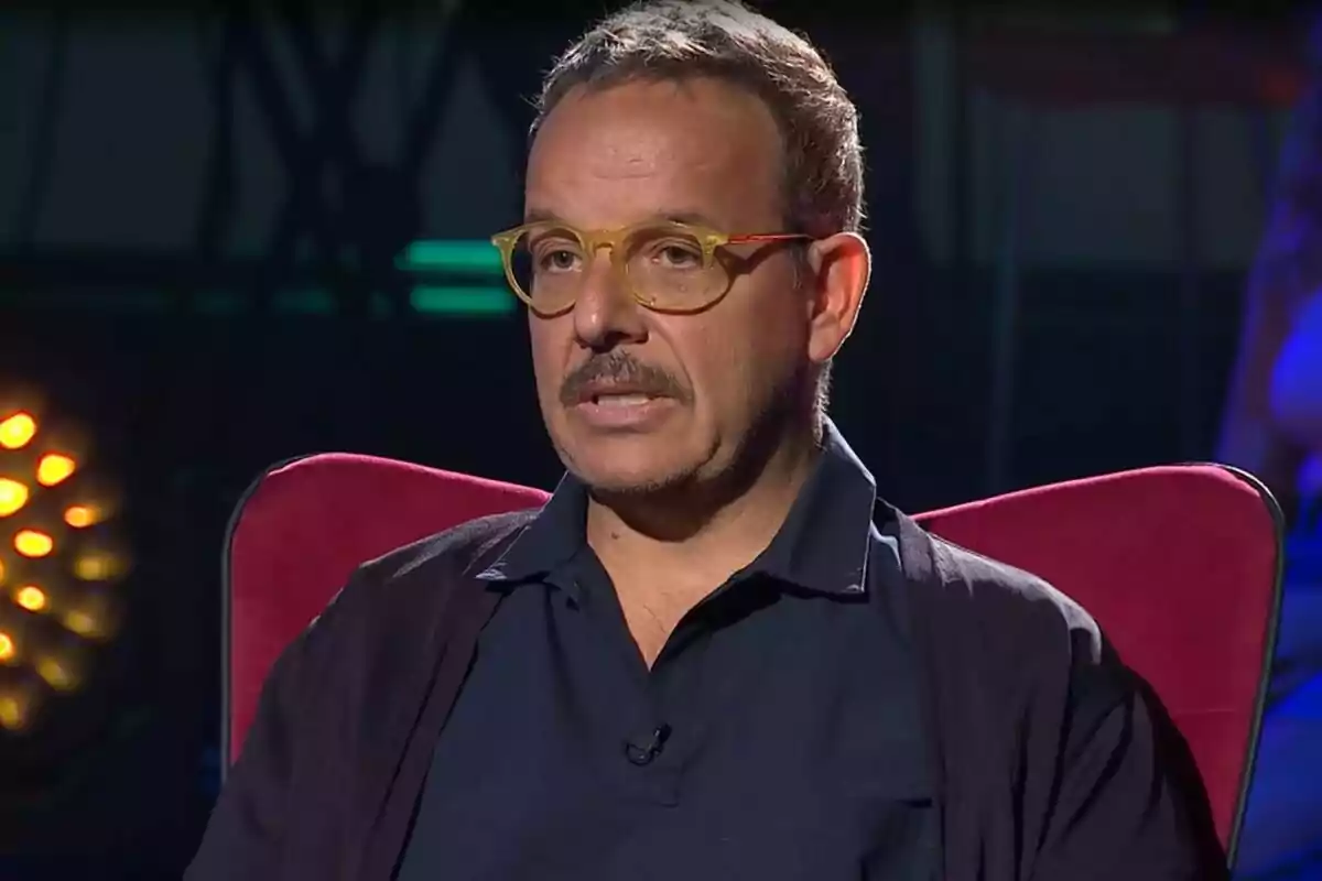 Hombre con gafas y bigote sentado en una silla roja en un entorno oscuro con luces de fondo.