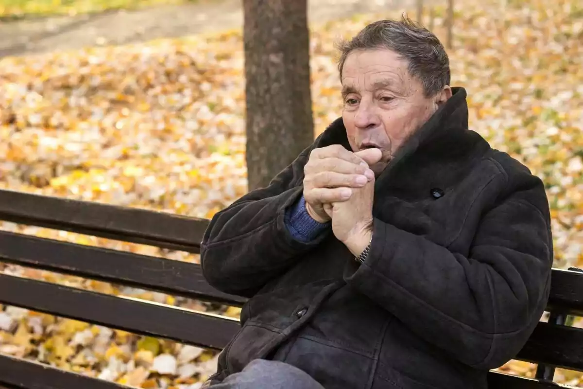 Hombre mayor sentado en un banco de parque en otoño calentándose las manos.