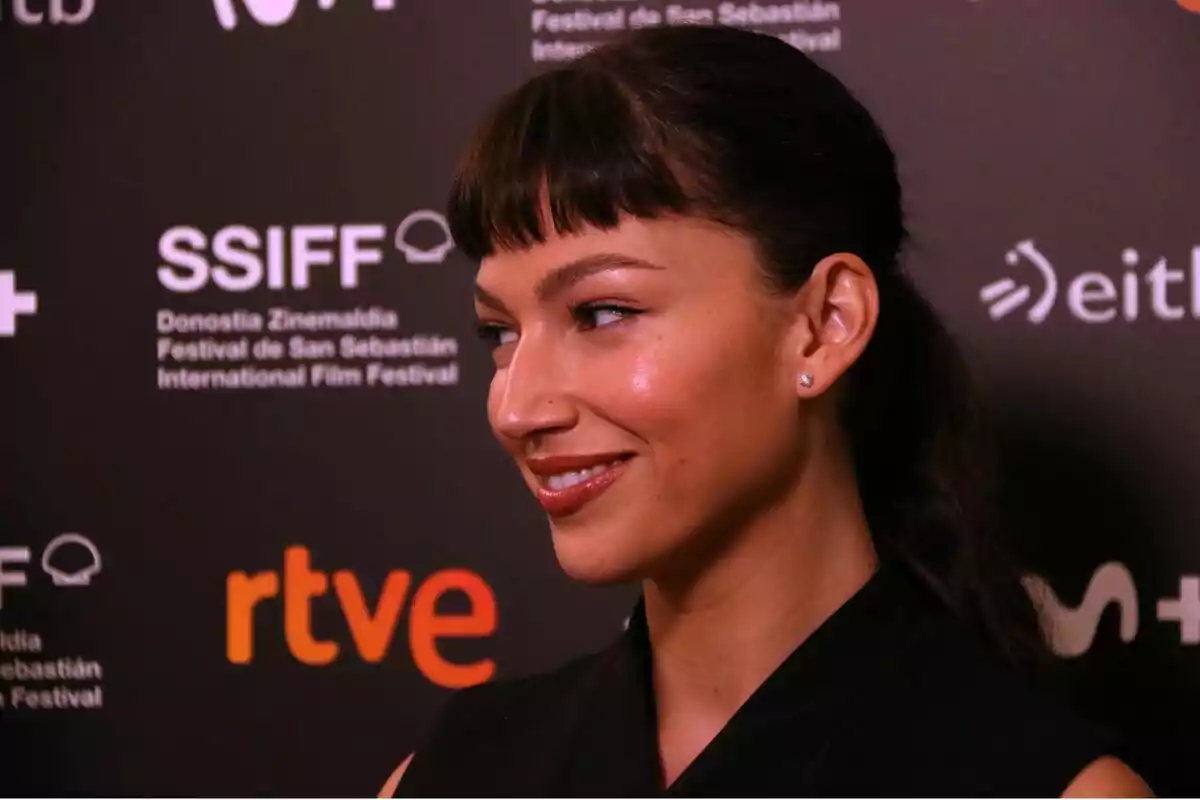 Una mujer sonriente con flequillo y cabello recogido, posando frente a un fondo con logotipos del Festival de San Sebastián y otras marcas.