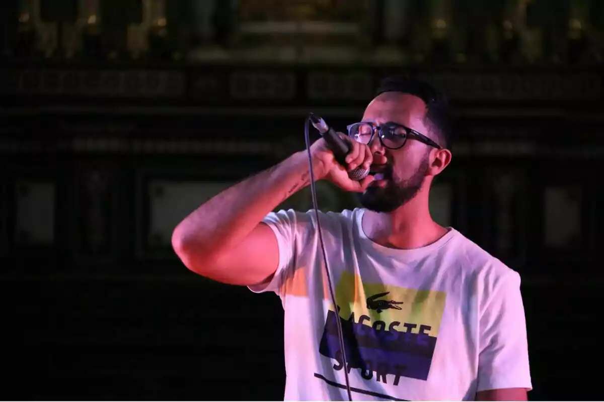 Un home amb ulleres i barba canta en un micròfon mentre fa servir una samarreta blanca amb el logo de Lacoste Sport.