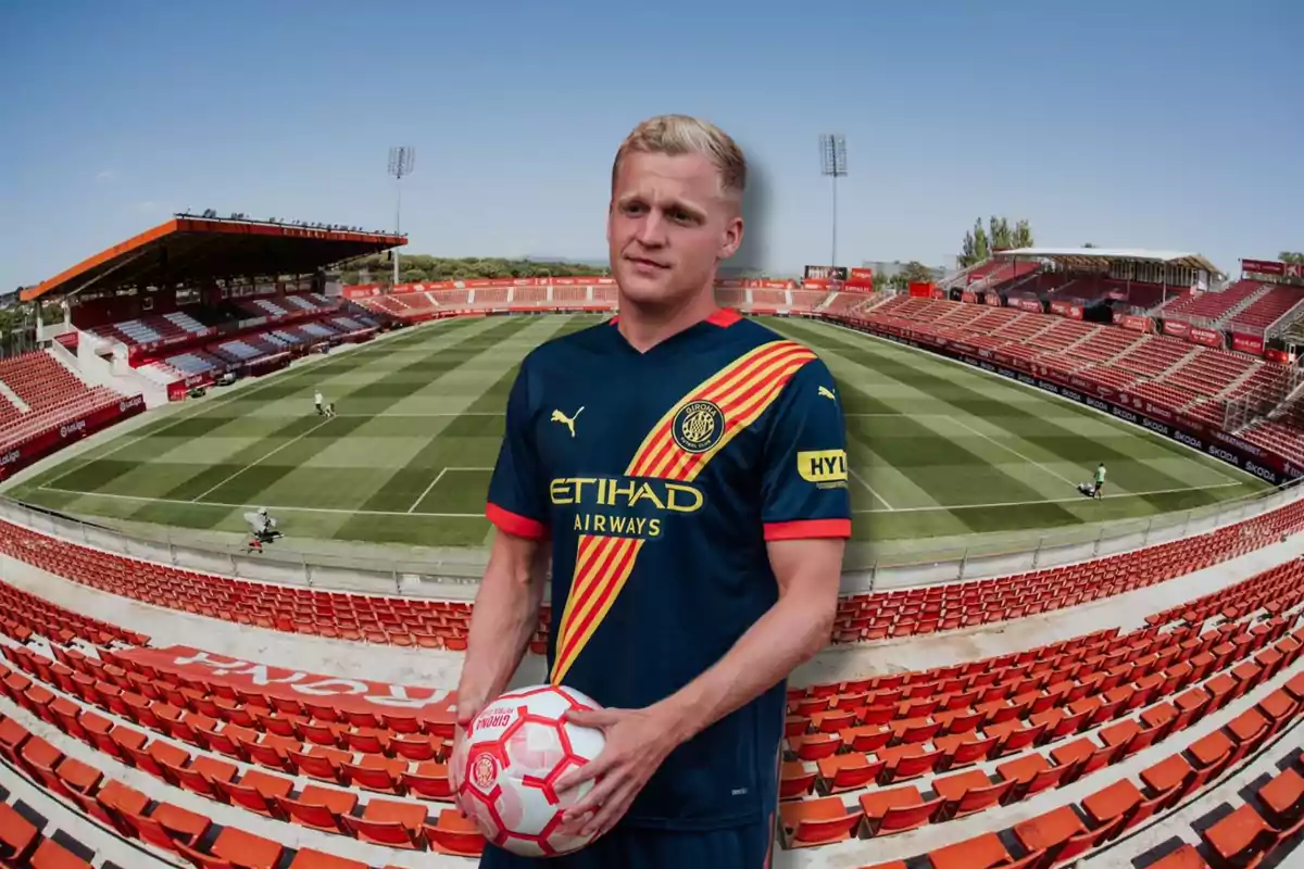 Un jugador de futbol amb uniforme blau fosc i detalls en groc i vermell sosté una pilota en un estadi buit amb seients vermells i un camp de gespa ben cuidat.