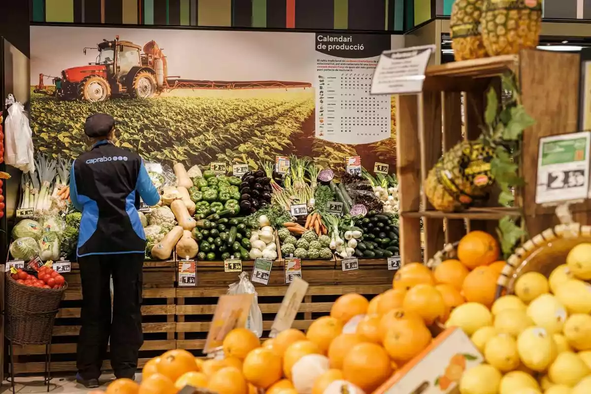 Un empleat de supermercat organitza verdures i fruites fresques a una secció de productes agrícoles, amb un fons que mostra un camp de cultiu i un calendari de producció.