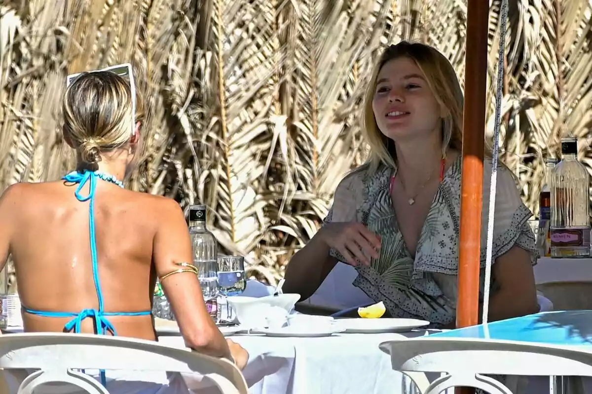 Victoria Iglesias y una amiga conversan mientras disfrutan de una comida al aire libre en un entorno tropical. Victoria Iglesias y una amiga conversan mientras disfrutan de una comida al aire libre en un entorno tropical.