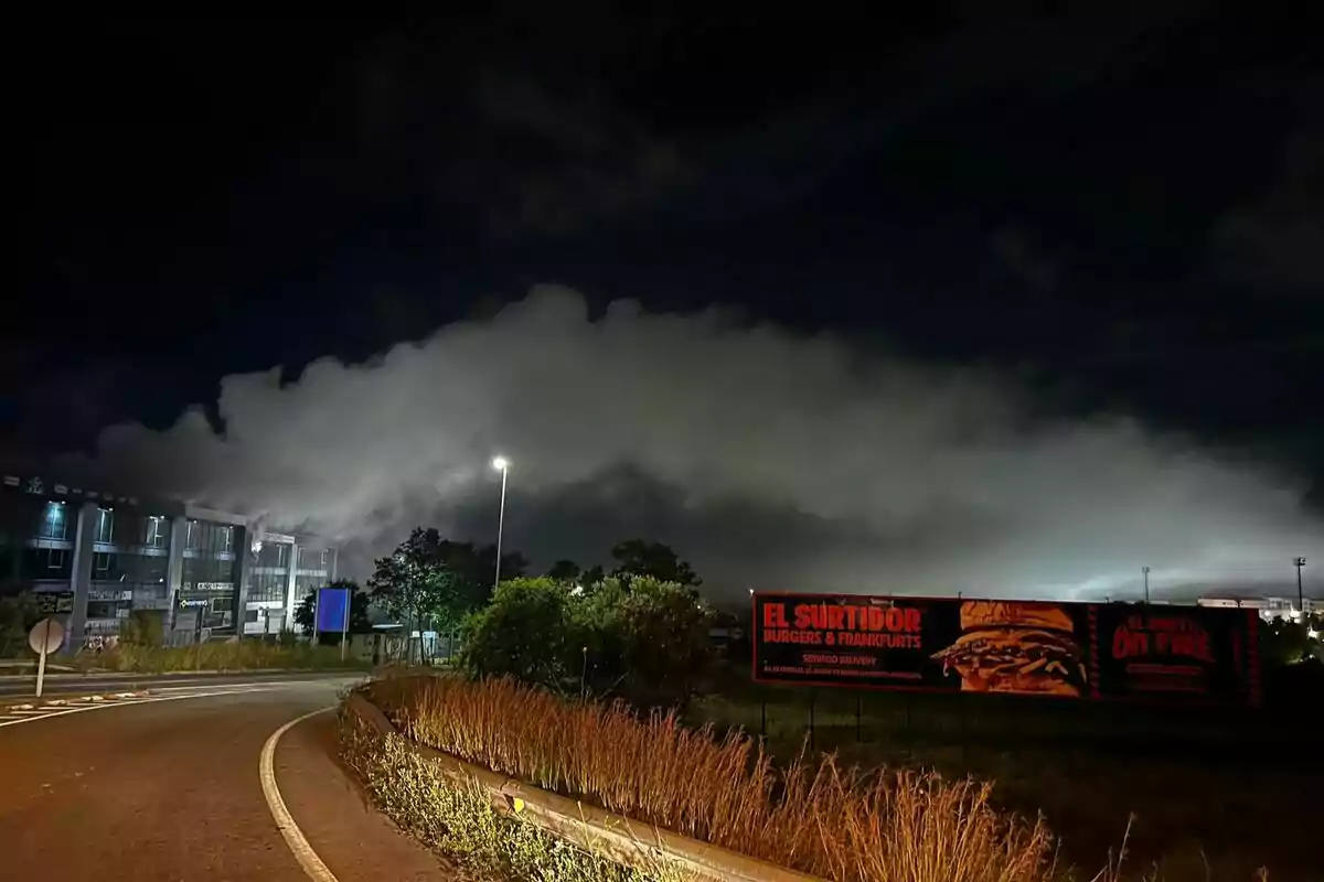 Una carretera iluminada por farolas en la noche con un edificio a la izquierda y un cartel publicitario de hamburguesas a la derecha, mientras una nube densa cubre el cielo. Una carretera iluminada por farolas en la noche con un edificio a la izquierda y un cartel publicitario de hamburguesas a la derecha, mientras una nube densa cubre el cielo.