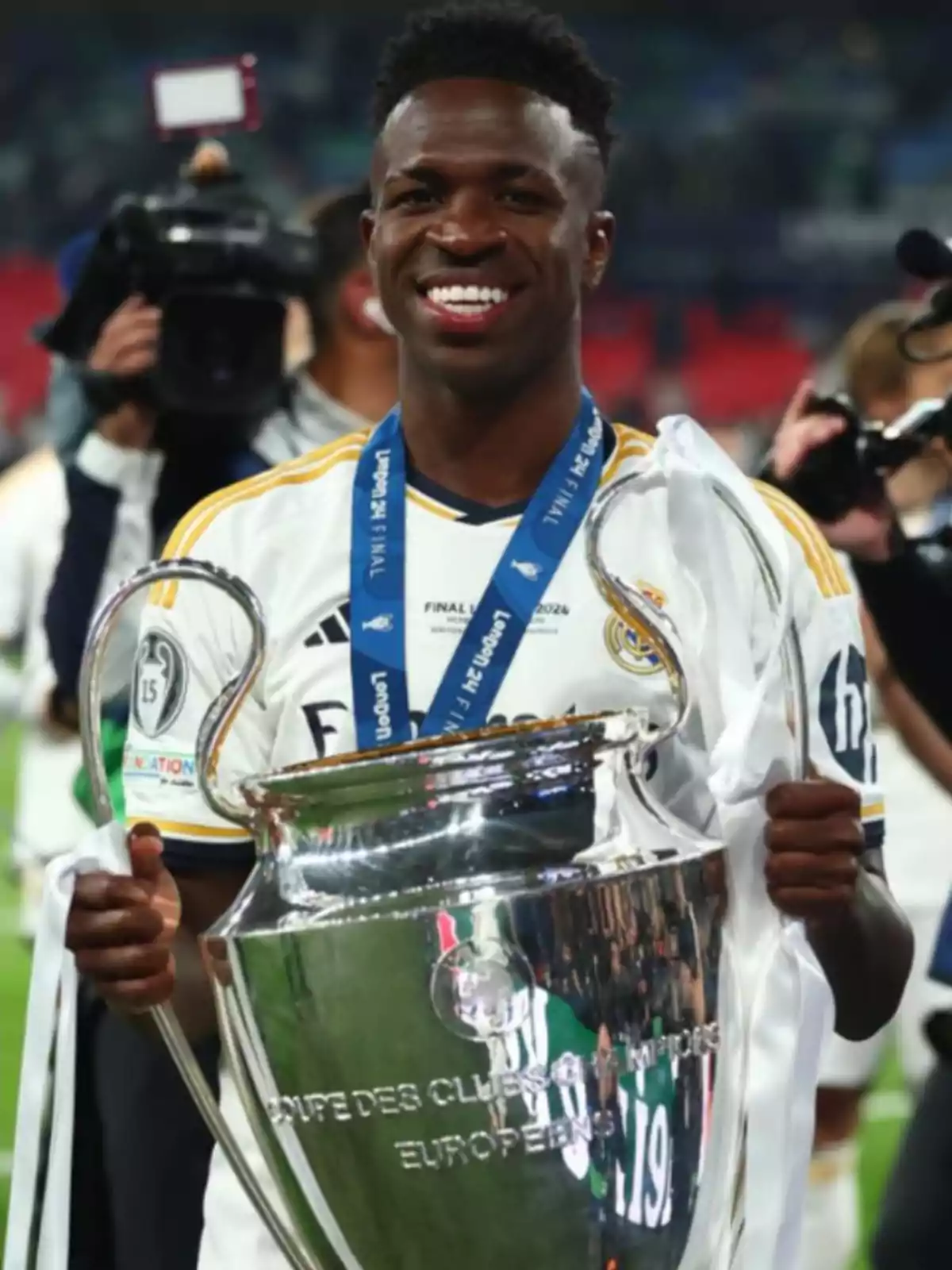 Un jugador de futbol somriu mentre sosté un trofeu gran i platejat, envoltat de càmeres i fotògrafs. Un jugador de futbol somriu mentre sosté un trofeu gran i platejat, envoltat de càmeres i fotògrafs.