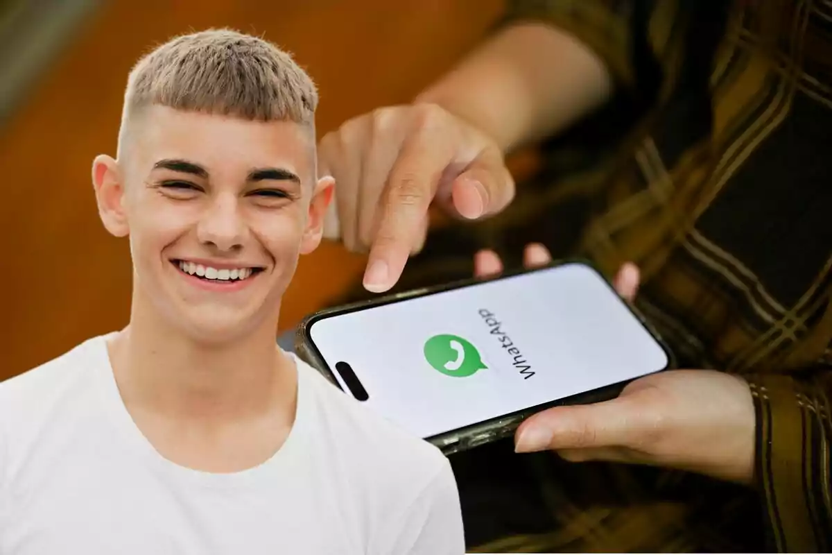 Un joven sonriente junto a una mano que sostiene un teléfono mostrando el logo de WhatsApp.