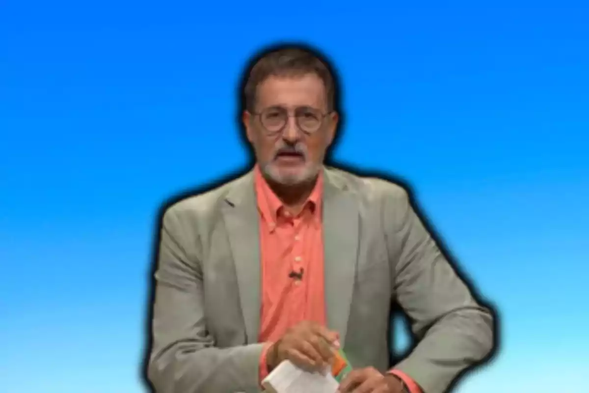 Un hombre con gafas y barba, vestido con saco verde y camisa naranja, sostiene un libro frente a un fondo azul.