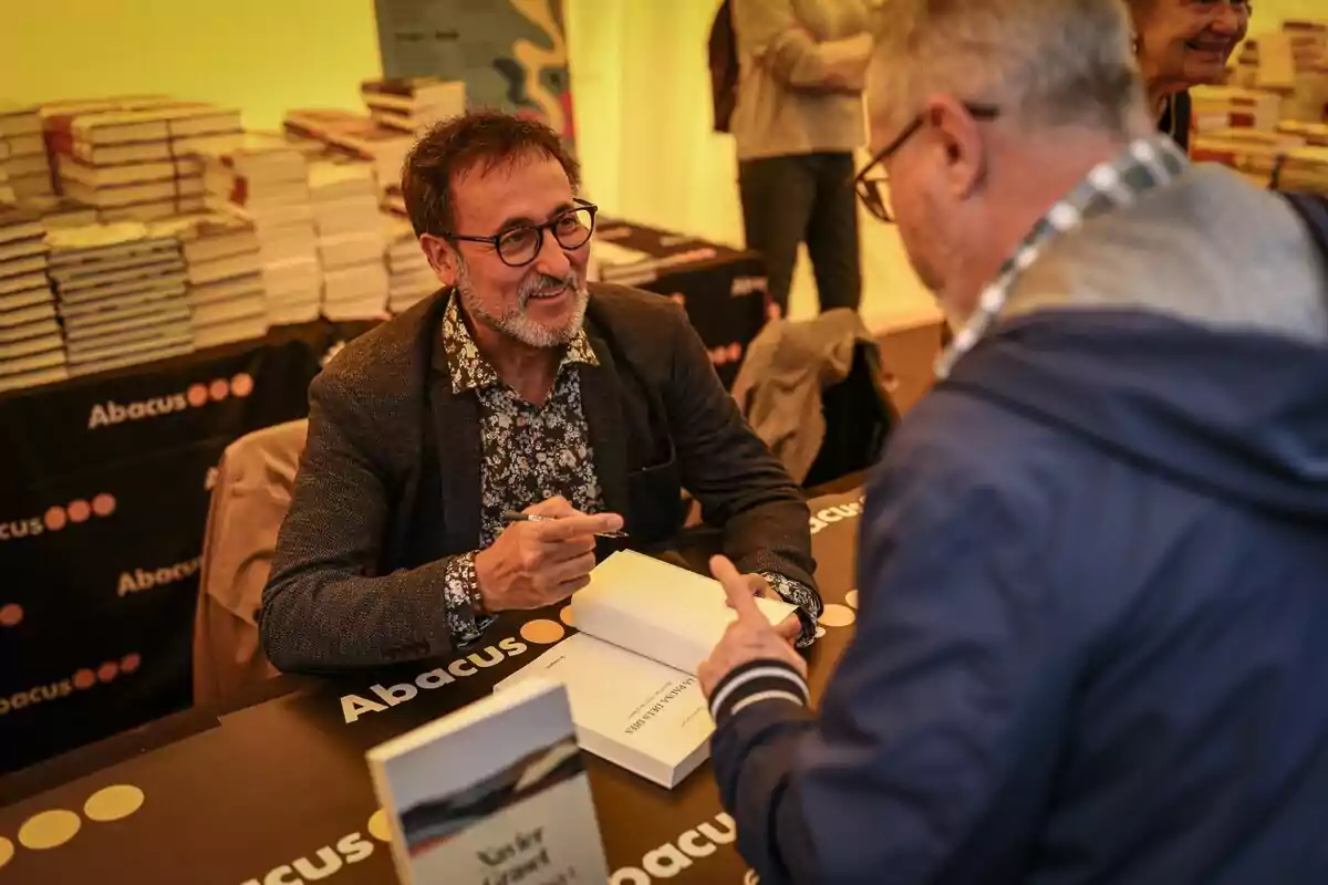 Un home amb ulleres i barba signa un llibre per a una altra persona en un esdeveniment d'Abacus, amb una pila de llibres al fons.