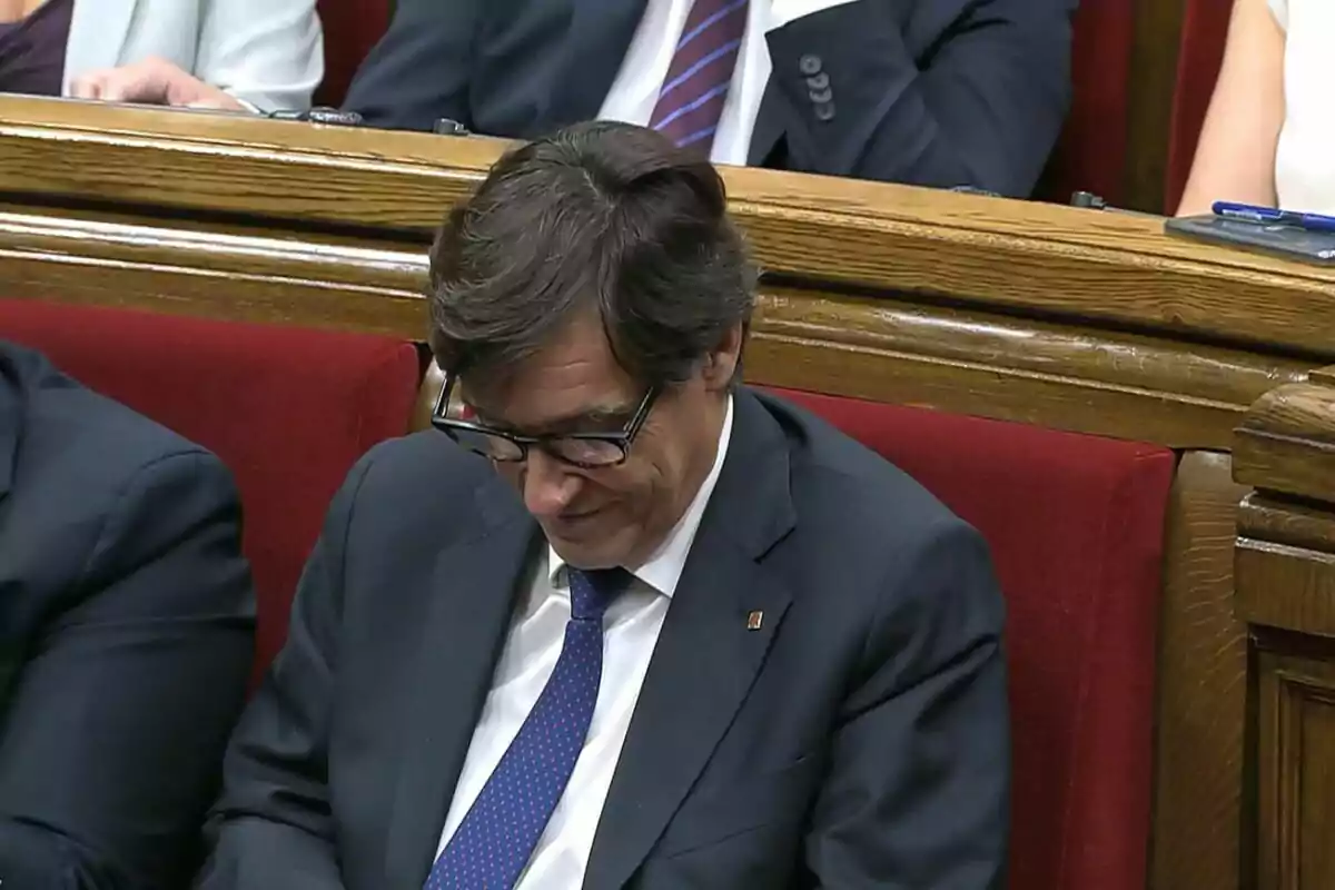 Un home amb ulleres i vestit fosc està assegut en un seient vermell dun parlament, mirant cap avall. Un home amb ulleres i vestit fosc està assegut en un seient vermell dun parlament, mirant cap avall.