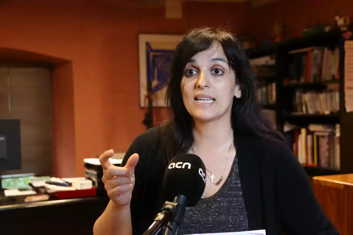 Dona parlant davant d'un micròfon en una habitació amb prestatgeries plenes de llibres. Dona parlant davant d'un micròfon en una habitació amb prestatgeries plenes de llibres.